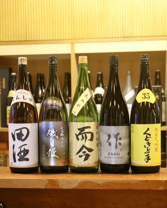 A shelf lined with 1.8-litre bottles of sake — Den-shu, Isojiman, Jikon, Zaku, Kudokijozu and more